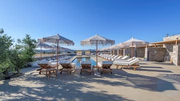 Zona junto a la piscina con sillones de descanso y sombrillas, cielo despejado y vista al mar.