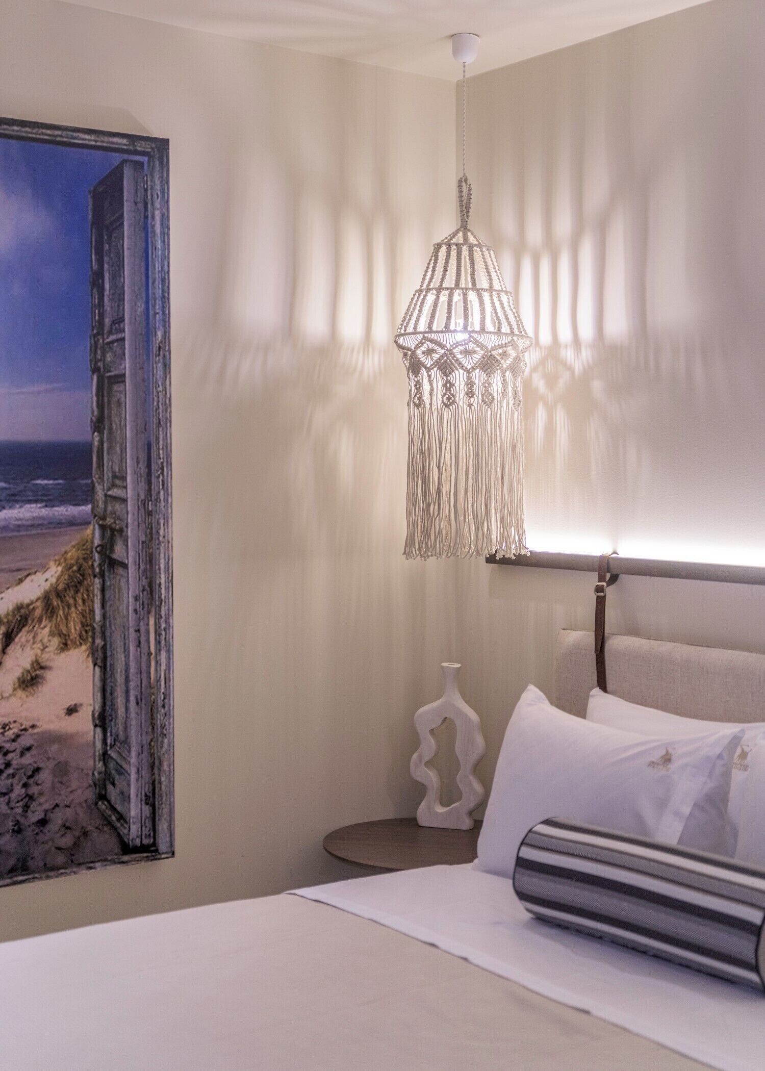 A bedroom with a bed, a hanging light fixture, a painting of a beach, and a bedside table with a vase.