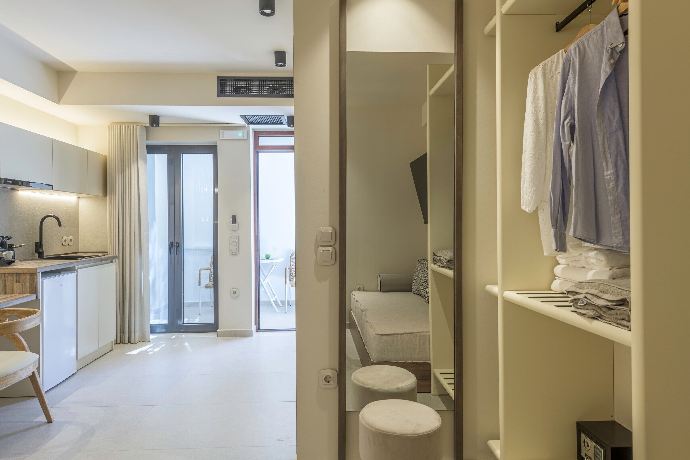 Ein modernes Hotelzimmer mit Küchenzeile, einem Bett mit weißem Kopfteil, einer kleinen Sitzecke und einem Badezimmer mit Handtuchhalter.