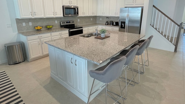 A modern kitchen with white cabinets, stainless steel appliances, a granite countertop, and a central island with bar stools. - 8957-HiltonRidge (Kissimmee)