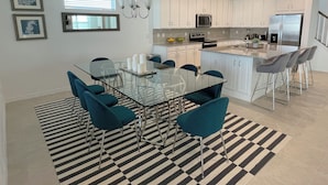 A modern kitchen with white cabinets, a glass dining table, teal chairs, and a striped rug. - 8957-HiltonRidge (Kissimmee)