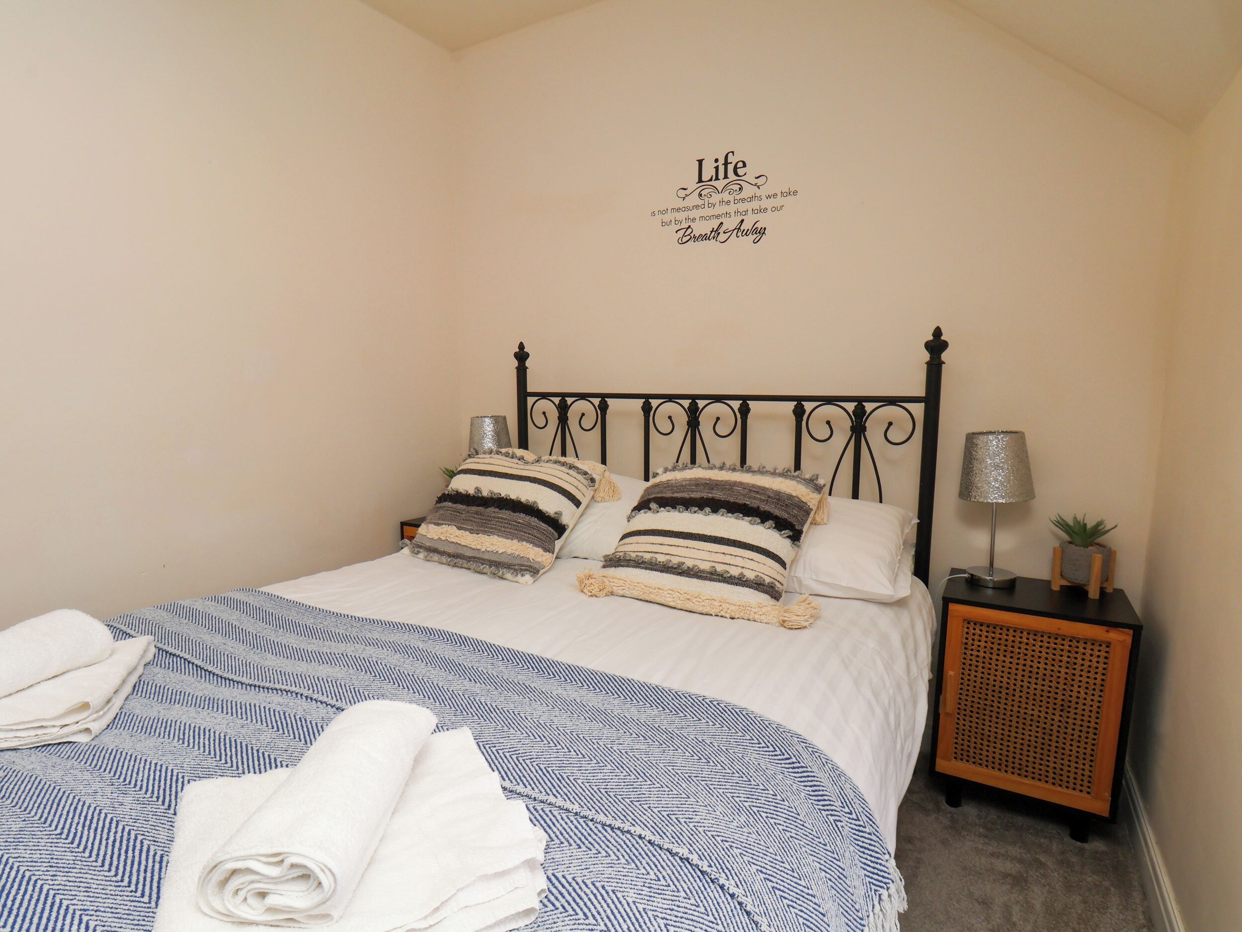 A neatly made bed with striped pillows, a wall quote, a bedside lamp, and a small plant on a nightstand.