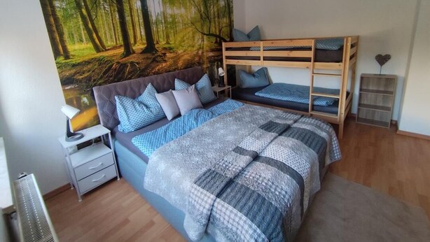 Une chambre avec un lit superposé, une grande fresque murale sur le thème de la forêt, une table de chevet avec une lampe et une petite table de nuit.