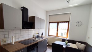 Une cuisine compacte équipée d’une cuisinière, d’un évier et de meubles de rangement modernes. On y trouve également un coin repas avec une table et des chaises, ainsi qu’une fenêtre avec des stores.