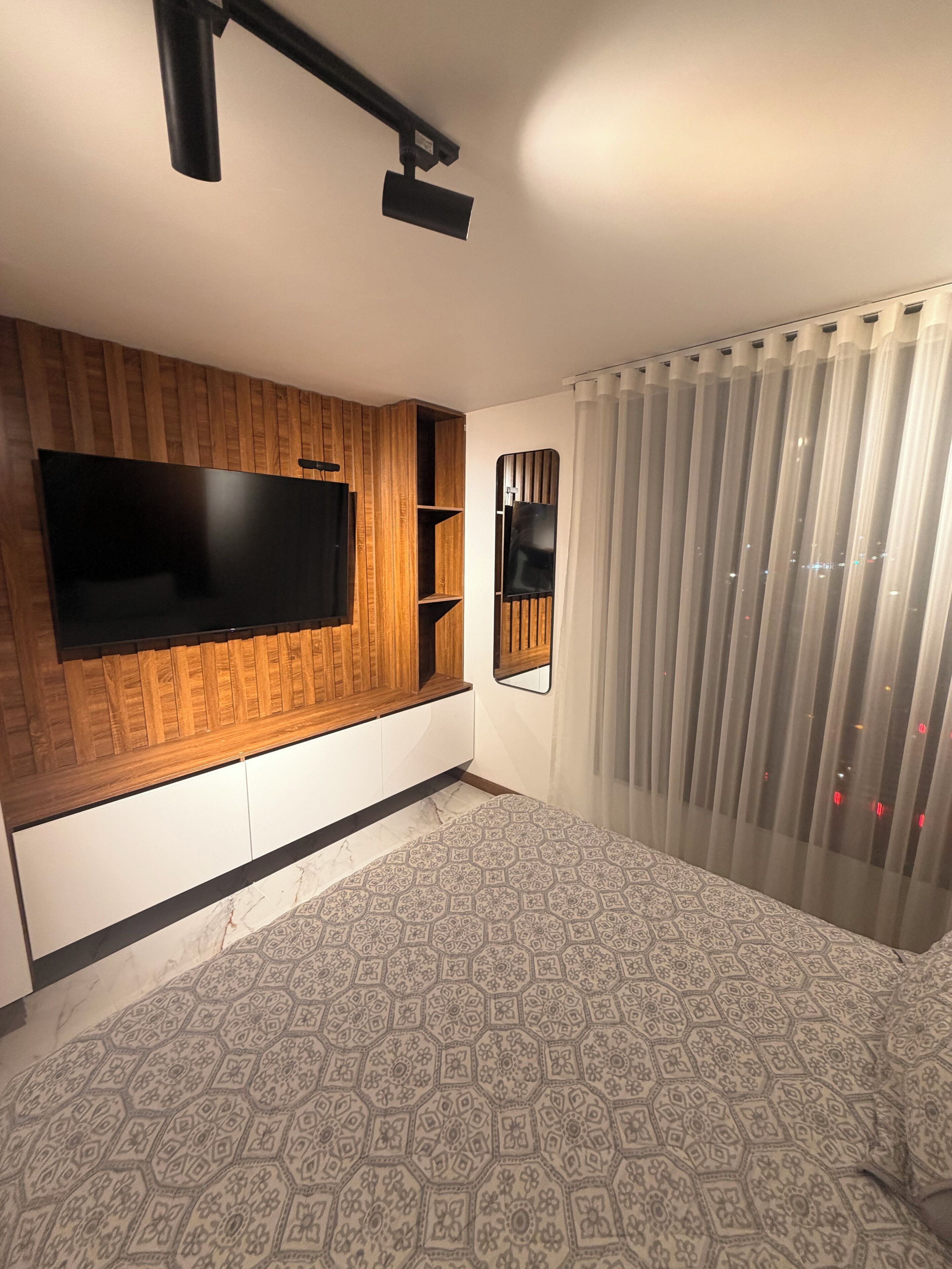 Un dormitorio con televisión instalada en una pared de paneles de madera, una cama con ropa de cama estampada y una ventana con cortinas traslúcidas.