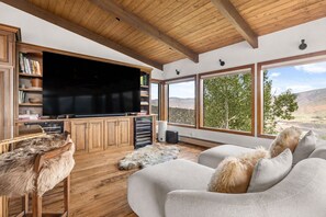 Ein modernes Wohnzimmer mit einem großen Flachbildfernseher, Holzkörperschränken, einem Fellteppich und Blick auf eine Berglandschaft durch große Fenster.