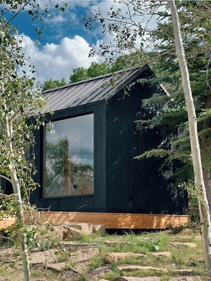 Eine kleine, dunkle Holzhütte mit einem großen Fenster, umgeben von Bäumen und Felsen.