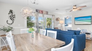 A modern living room with a wooden dining table, white chairs, a blue sofa, a chandelier, and a television. - Beachside Villas 1023 (Santa Rosa Beach)