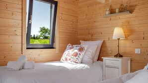 Ein Zimmer mit Holzwänden, einem Bett, einem Nachttisch, einer Lampe und einem Fenster mit Blick auf Grünfläche.