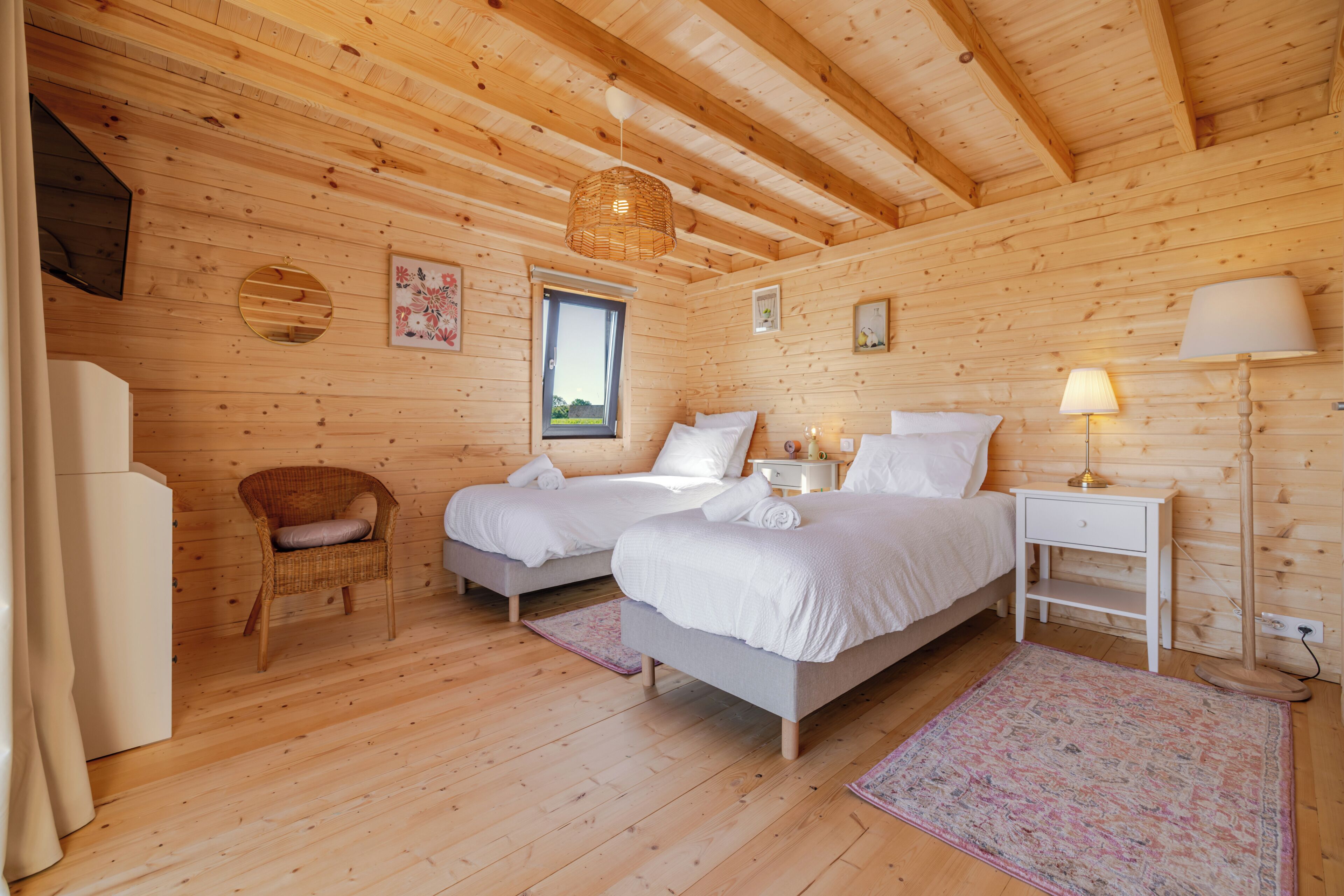 Una habitación de cabaña de madera con dos camas, una silla, una mesita de noche, una lámpara y una ventana.