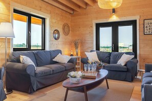Una habitación acogedora con dos sofás grises, piso de madera, una mesa de centro redonda de madera y un gran ventanal con vista a una playa.