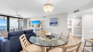 A modern living room with a glass-top dining table, a blue sofa, wooden chairs, and a chandelier. - Watercrest 1006 (Panama City Beach)