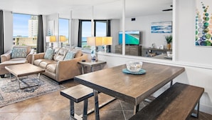 Un salon moderne comprenant un canapé, une table basse et une table à manger, de grandes fenêtres offrant une vue sur la ville et un téléviseur à écran plat.