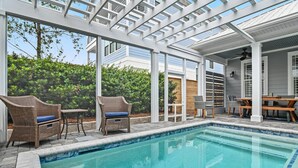 Zona piscina con una pergola, mobili in vimini e vista su un edificio residenziale.