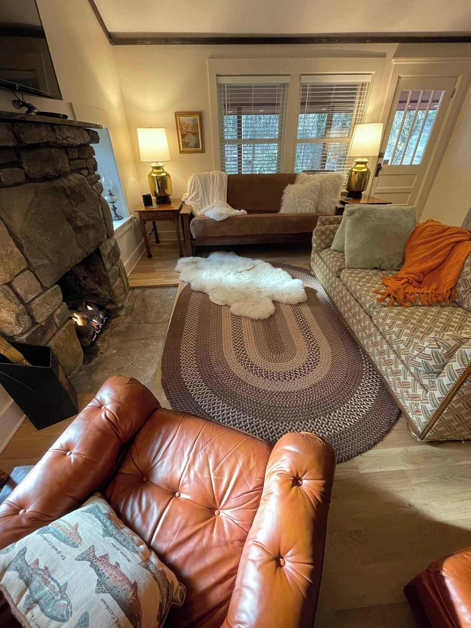 En hyggelig stue med en stenspejs, en læderarmstol, en stribet sofa og en dekorativ måtte.