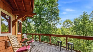 A wooden deck with a rocking chair and a table, overlooking a forested area. - Luxury Treehouse, gas fireplace, BBQ, firepit, outdoor dining, shared creek access (Weaverville)
