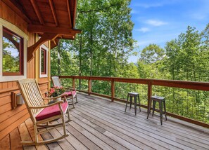 Eine Holzterrasse mit Schaukelstuhl und Tisch, mit Blick auf ein bewaldetes Gebiet.