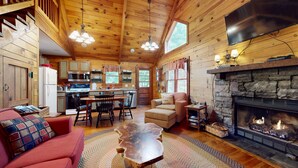 L’intérieur d’un chalet accueillant, avec une cheminée en pierre, un plafond en bois et un coin repas donnant sur une cuisine en arrière-plan.