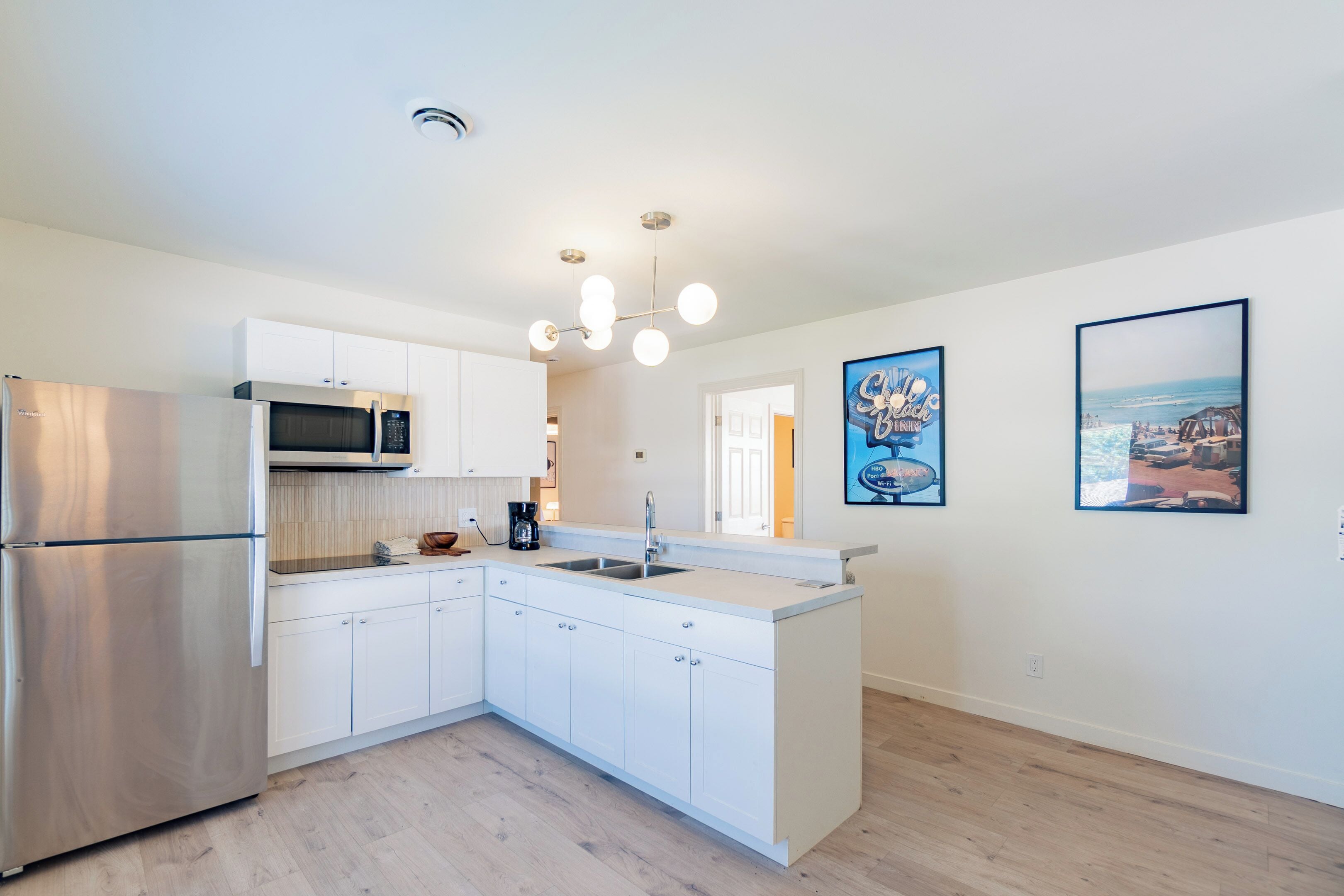 A modern kitchen with white cabinetry, a stainless steel refrigerator, a microwave, and a sink. There are two framed pictures on the wall.