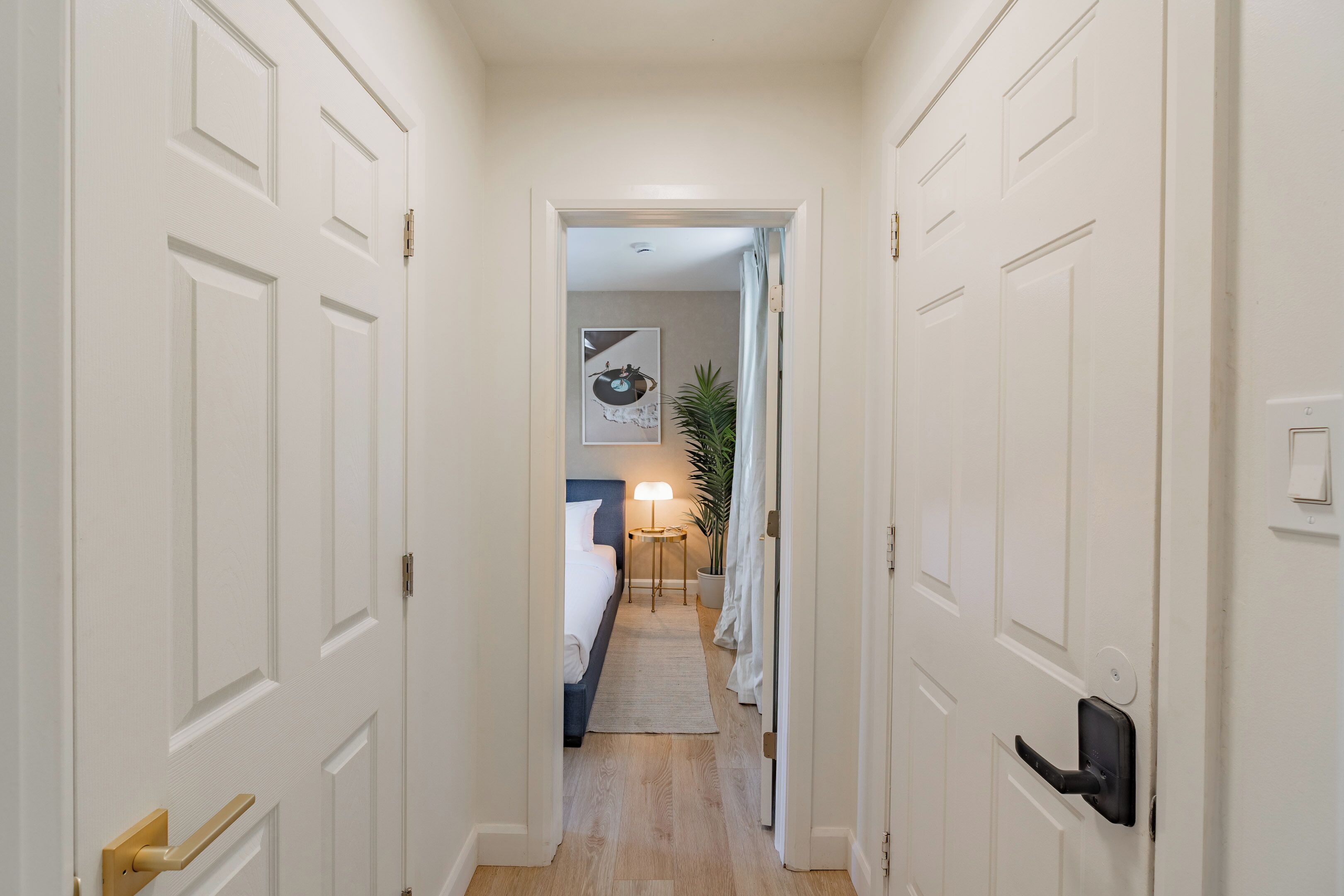 Un pasillo que conduce a un dormitorio con cama, mesita de noche, lámpara y planta en maceta.