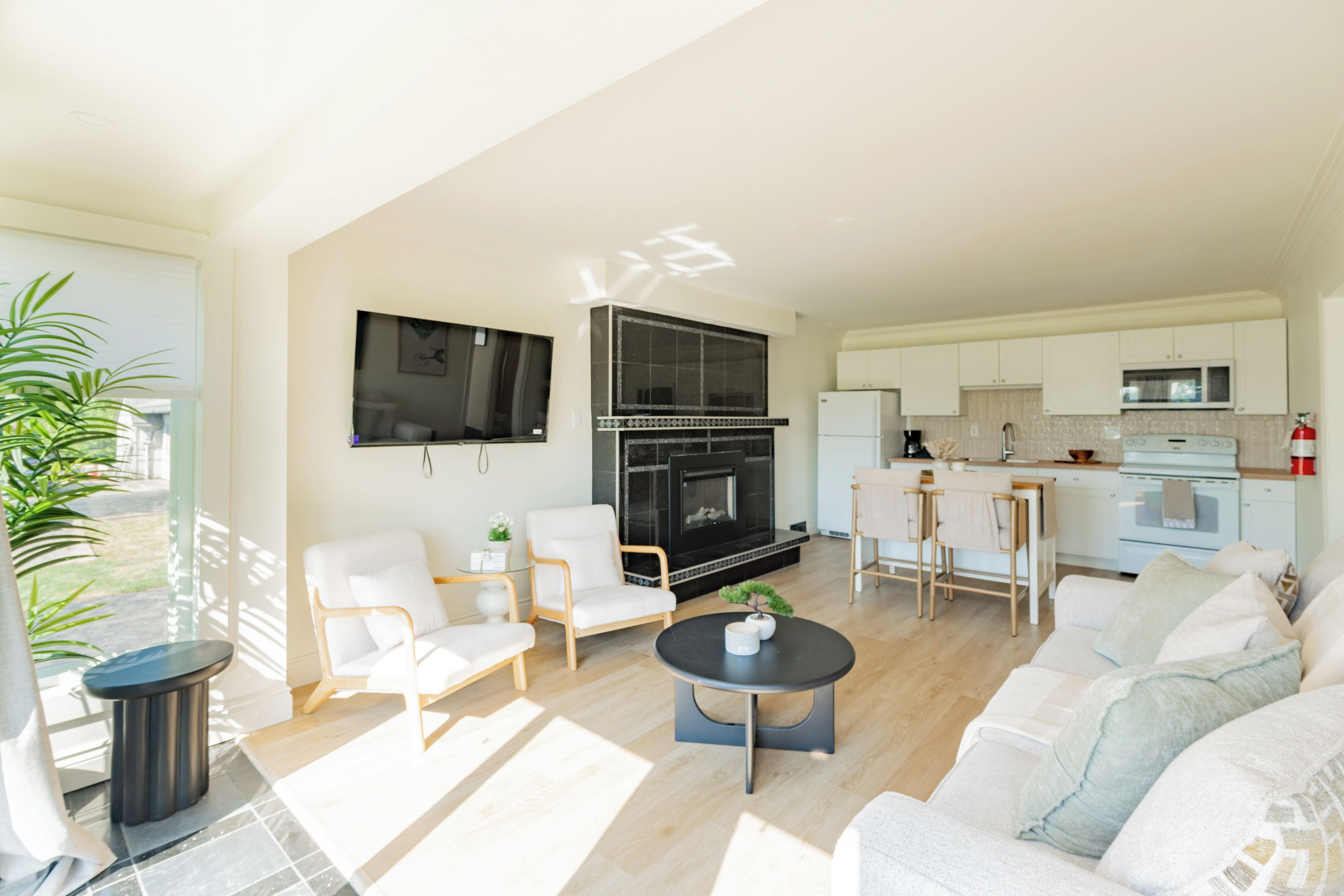 A modern living room with a fireplace, white sofas, a coffee table, and a kitchen area with white cabinets and appliances.