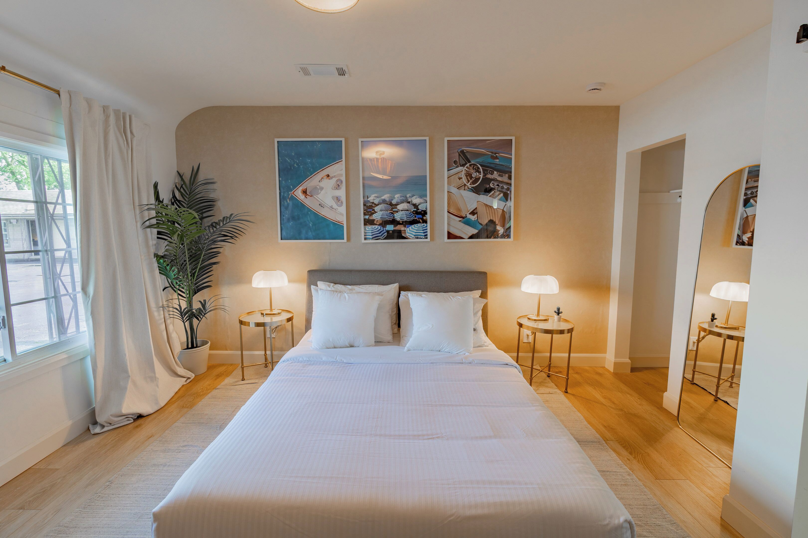 A large bed with white linens, two bedside lamps, a potted plant, and three framed pictures on the wall.