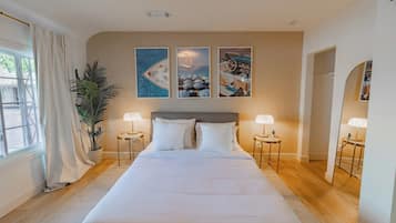 A large bed with white linens, two bedside lamps, a potted plant, and three framed pictures on the wall.