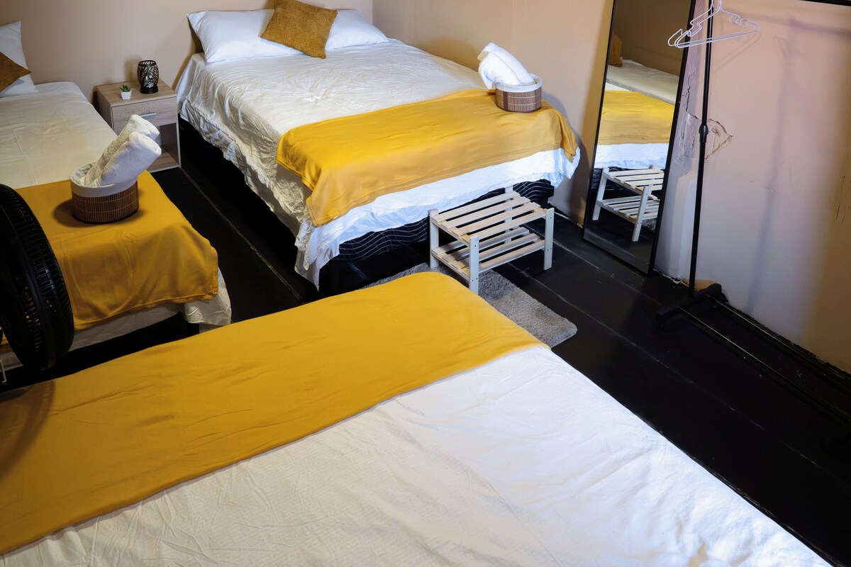 A hotel room with two beds, each with a yellow bedspread, a small wooden stool, and a mirror on the wall.
