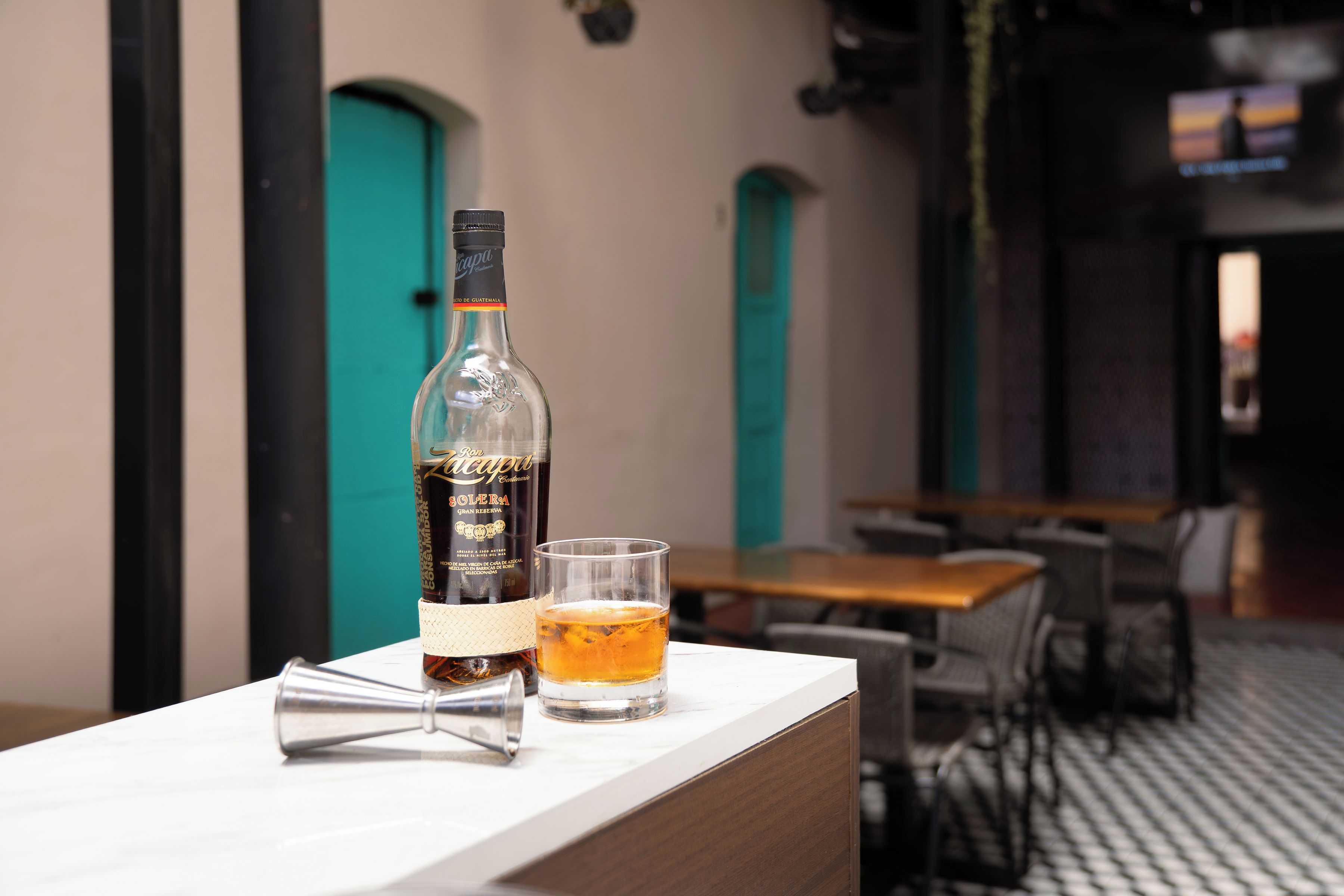 A bottle of Ron Zacapa Solera Gran Reserva on a table with a glass of amber liquid, a cocktail shaker, and a bar area in the background.