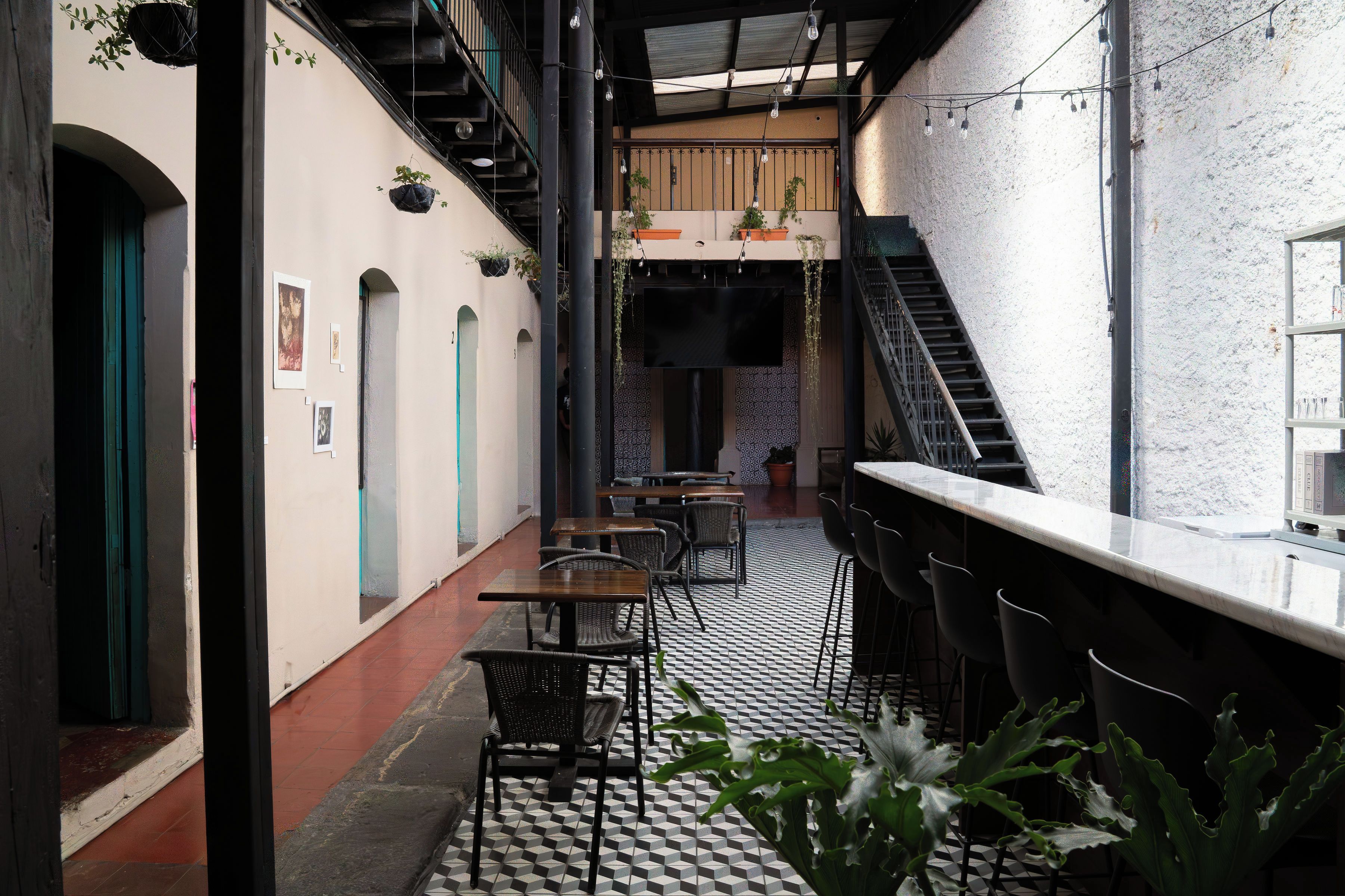 A narrow corridor with a bar area on one side, featuring a checkered floor, black metal chairs, and a staircase leading to an upper level.