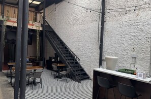 Un área de estar al aire libre con una escalera que conduce a un nivel superior, rodeada por una pared con textura y luces colgantes.
