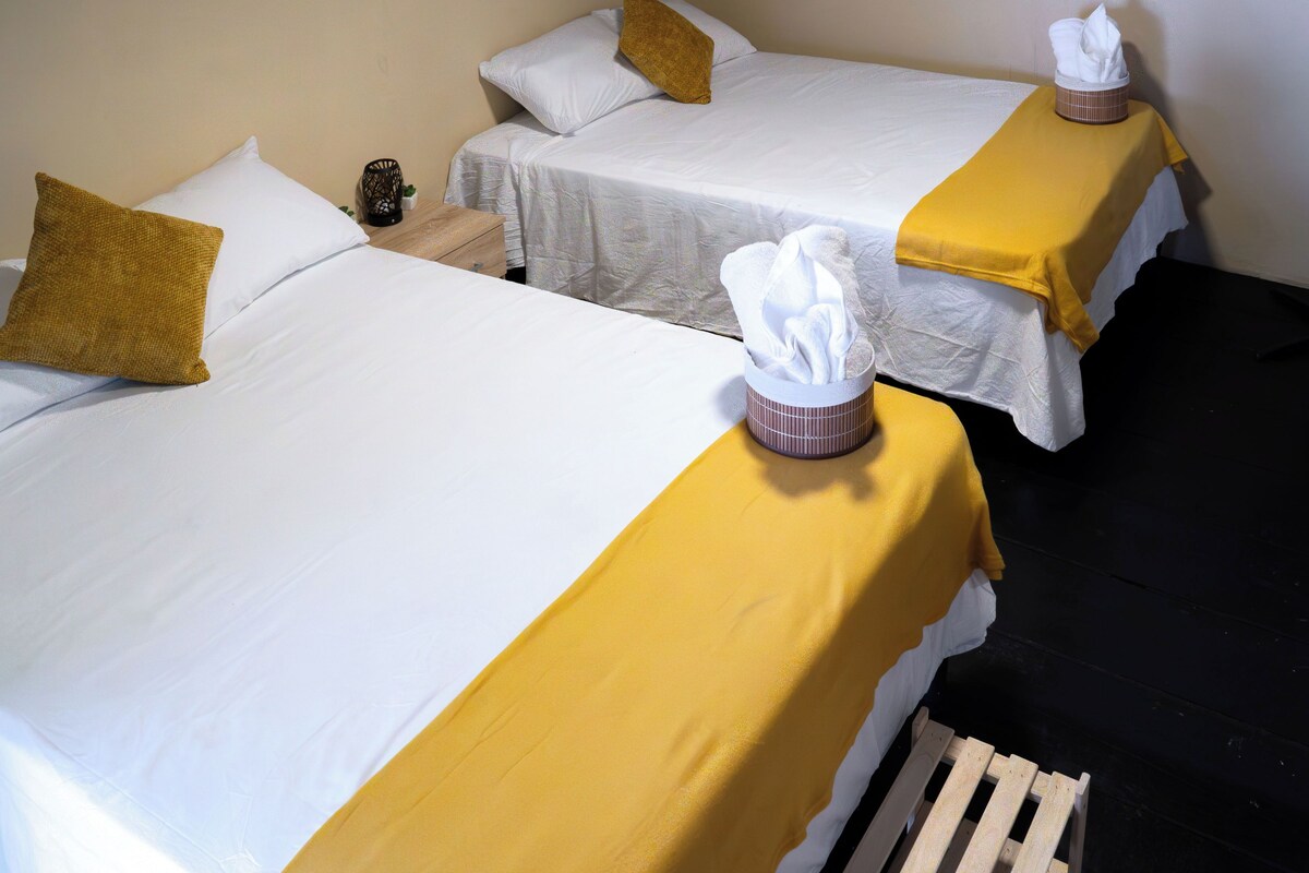 A hotel room with two beds, each with a white bedspread and a yellow accent. There are two white pillows and a brown accent pillow on each bed. A tissue box is placed on the bedside table.