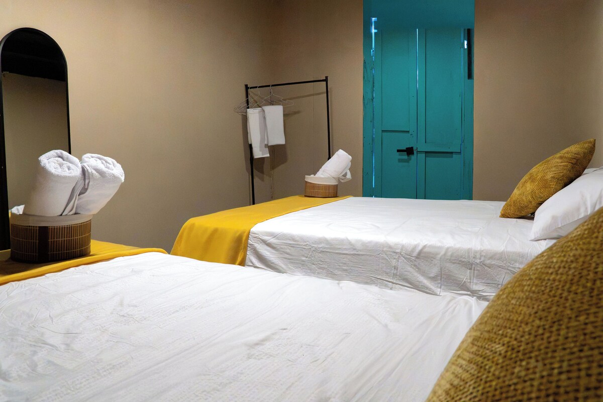 A hotel room with two beds, a mirror, a towel rack, and a teal door.