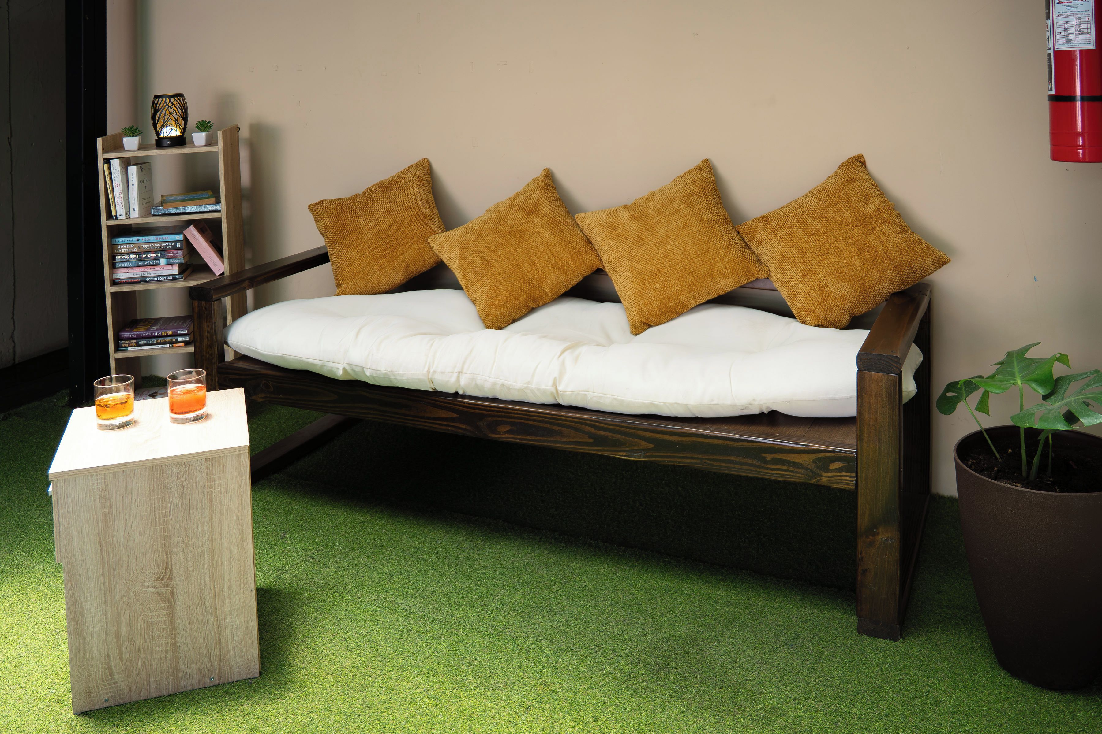Una habitación con un sofá de madera con cojines blancos, una mesita de madera con dos vasos, una estantería con libros y una planta en maceta.