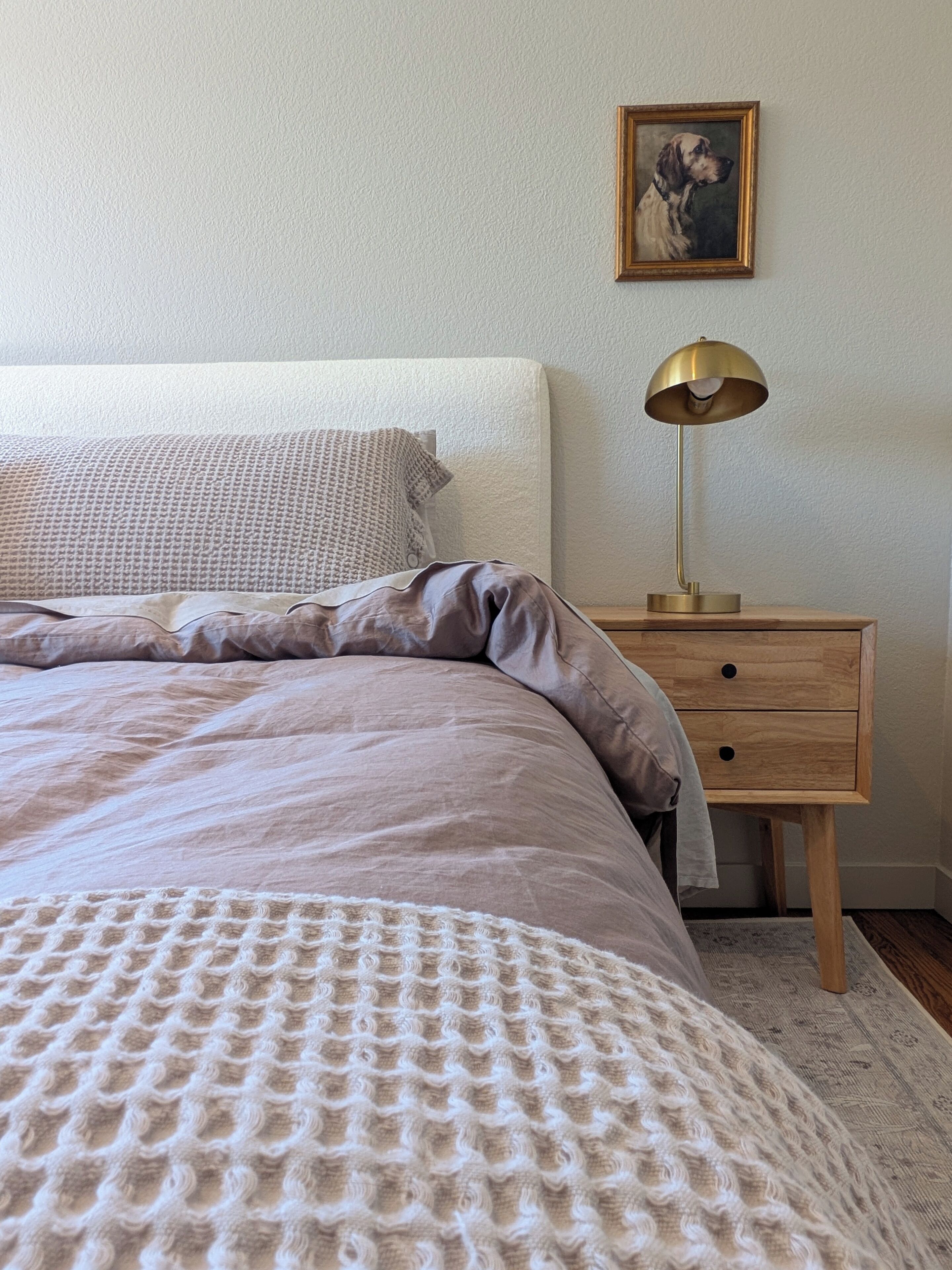 A neatly made bed with a textured blanket, a bedside lamp, a wooden nightstand, and a framed picture on the wall.