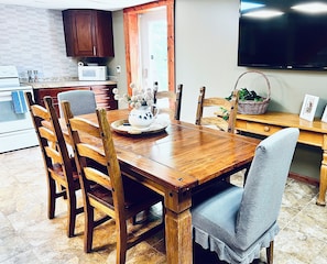 Una mesa de comedor de madera con sillas, un área de cocina con microondas y un televisor en la pared.