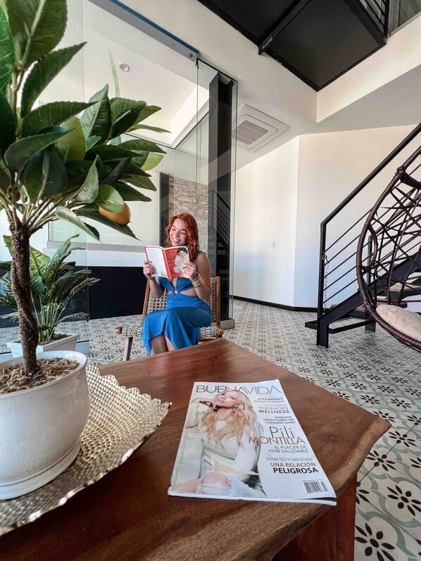 Una mujer leyendo una revista en una habitación moderna con una escalera y plantas en macetas.