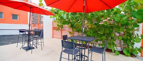 An outdoor patio with tables and chairs under red umbrellas, surrounded by greenery and orange buildings.
