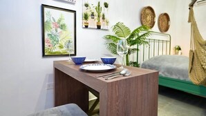 A small room with a bed, a hammock, a dining table with plates and cutlery, and wall art including a framed botanical illustration and potted plants.