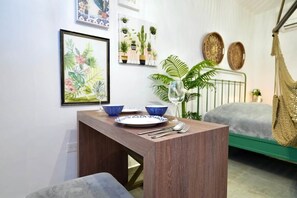 Una habitación pequeña con cama, una hamaca, una mesa de comedor con platos y cubiertos, y detalles decorativos como una ilustración botánica enmarcada y plantas en macetas.