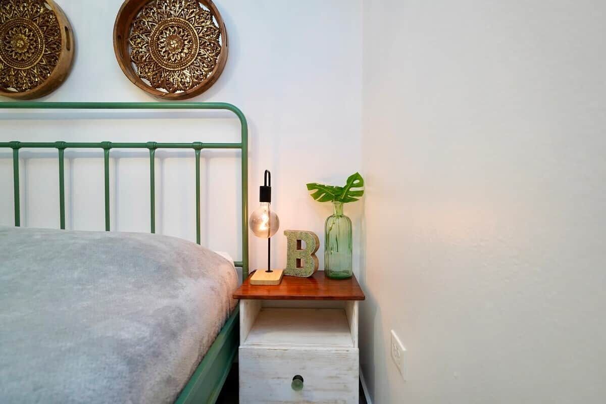 Ein Bett mit grünem Metallgestell, ein Nachttisch mit Lampe, ein Buchstabe 'B' und eine Pflanze.