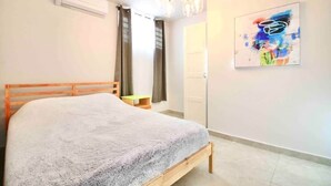 A bedroom with a bed, an air conditioner, a chandelier, a door, and a painting on the wall.