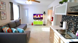 A modern living room with a sofa, a television, a ceiling fan, and a kitchen area with a sink and cabinets.