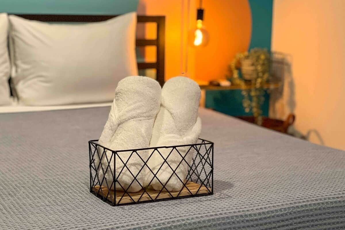 Una cama con un cojín blanco y una cesta de mimbre negra que contiene dos toallas blancas.
