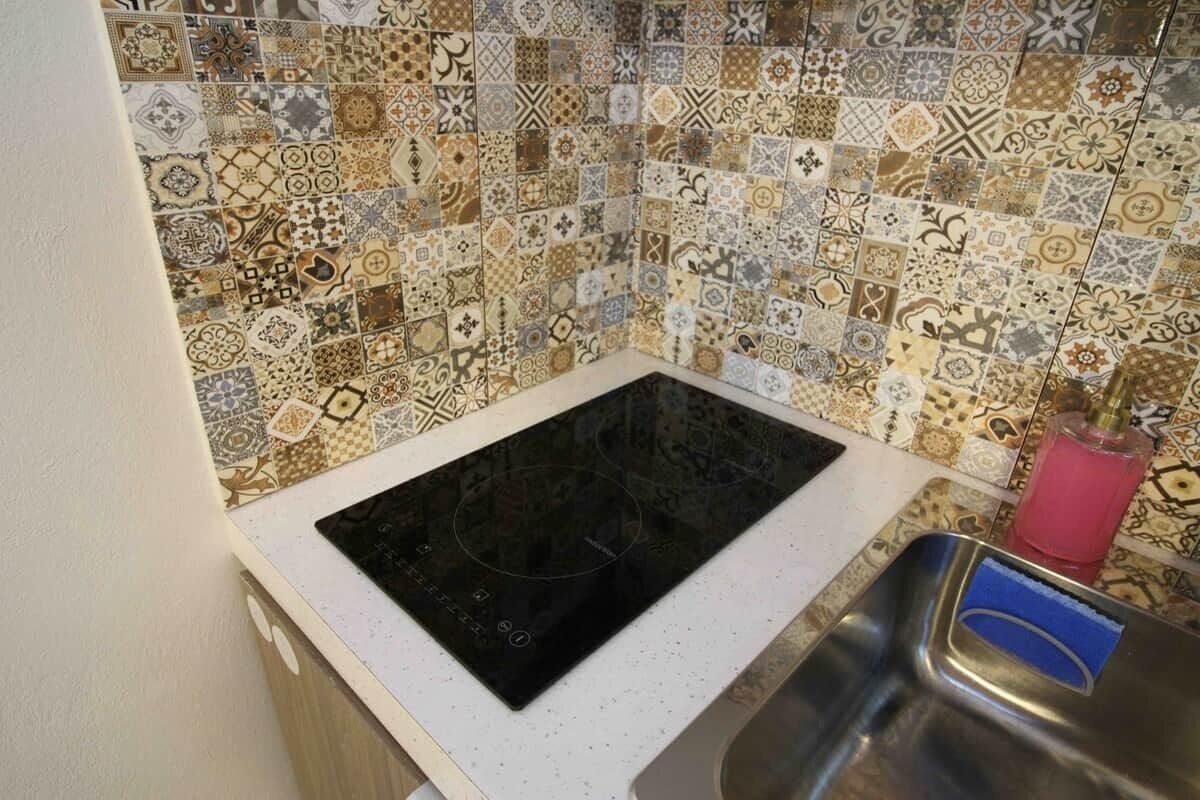 A kitchen with a black induction cooktop, a stainless steel sink, and a tiled backsplash with various patterns.