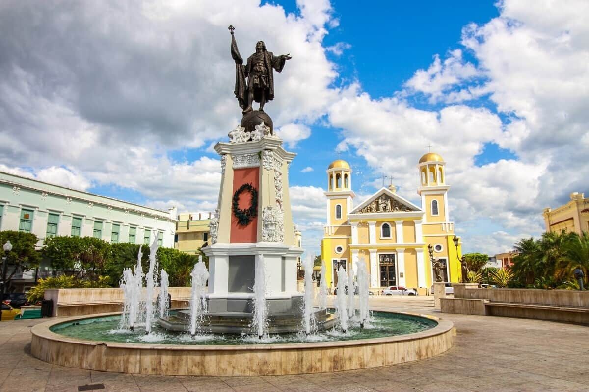 Una fuente con una estatua, rodeada por una plaza con edificios al fondo.