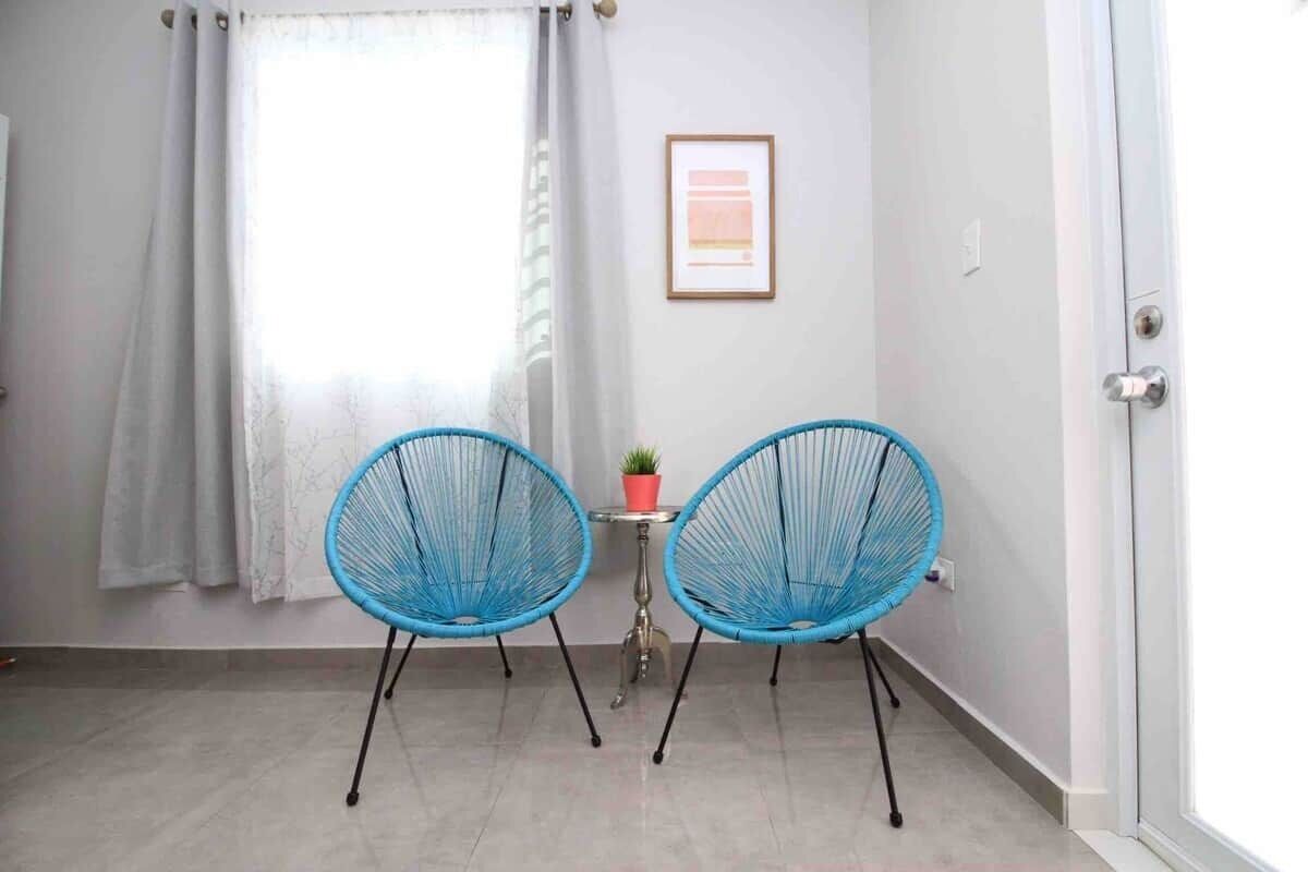 Una habitación con dos sillas azules, una mesita con una planta en maceta, un cuadro en la pared y una ventana con cortinas.