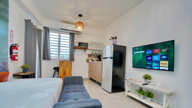 A modern hotel room with a bed, seating area, kitchenette, and a TV displaying various channels.