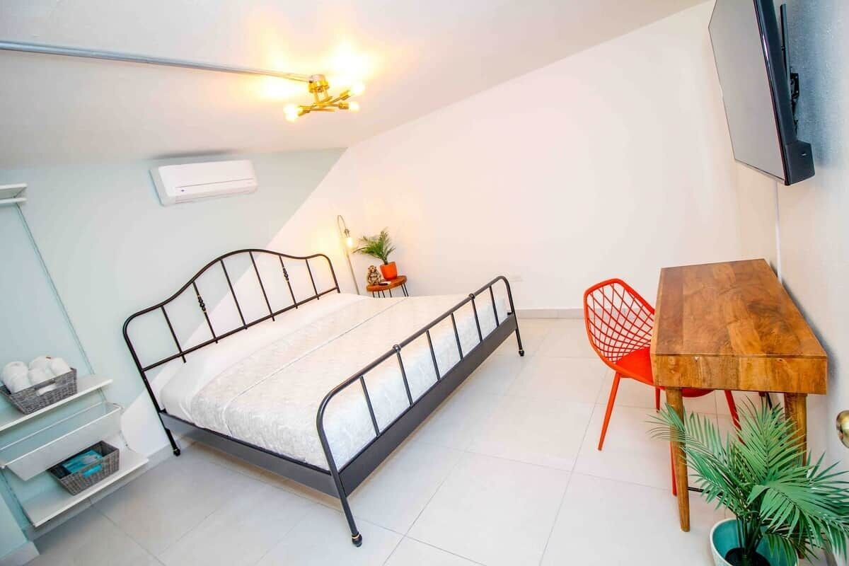 A bedroom with a bed, a desk, a chair, a potted plant, and a wall-mounted TV.