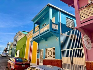 A street with colorful buildings, a car parked on the side, and a gated entrance. - 5.8 Casita Azul • Balcony • Terrace • Urban (Mayagüez)